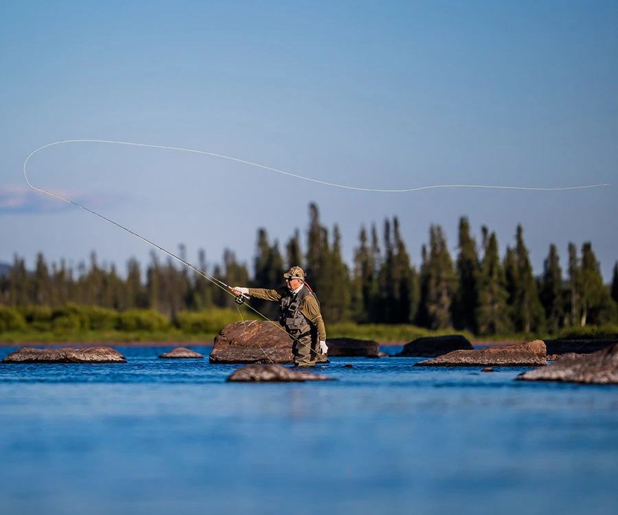 An angler in waders stands among rocks in a wide, shallow river and casts a long loop of fly line, with spruce forest and a blue sky behind.