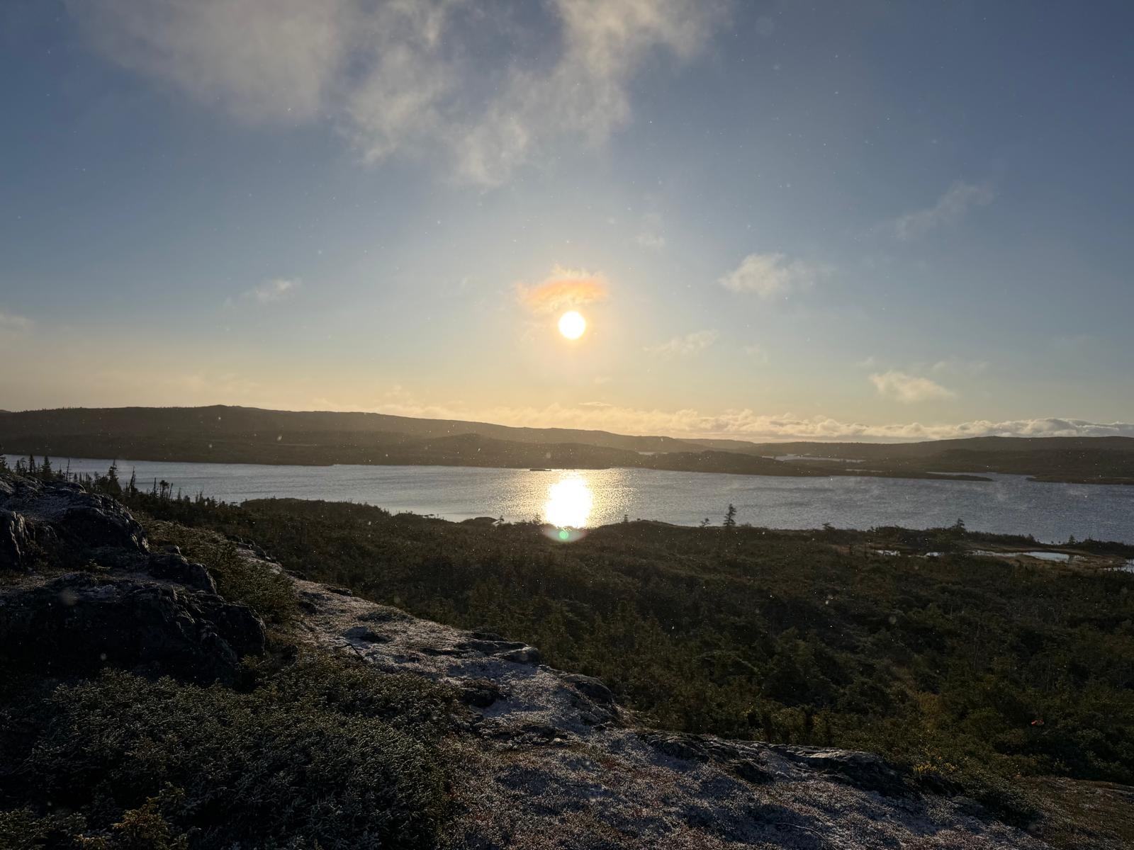 The sun hangs low over a lake and distant hills, its reflection on the water, with a rocky shore in front.