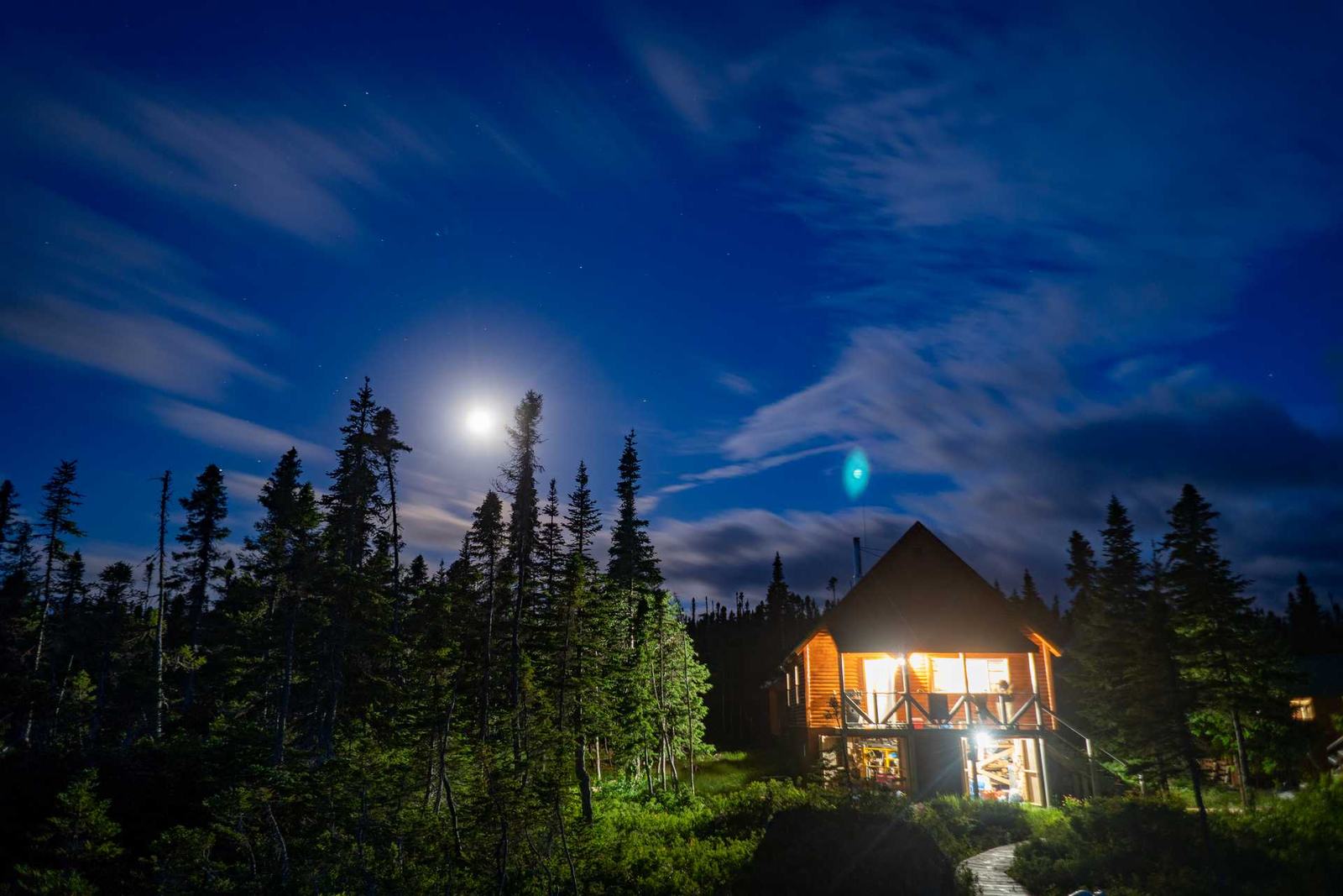 A lodge glows with warm light among spruce under a bright moon and a deep blue night sky.