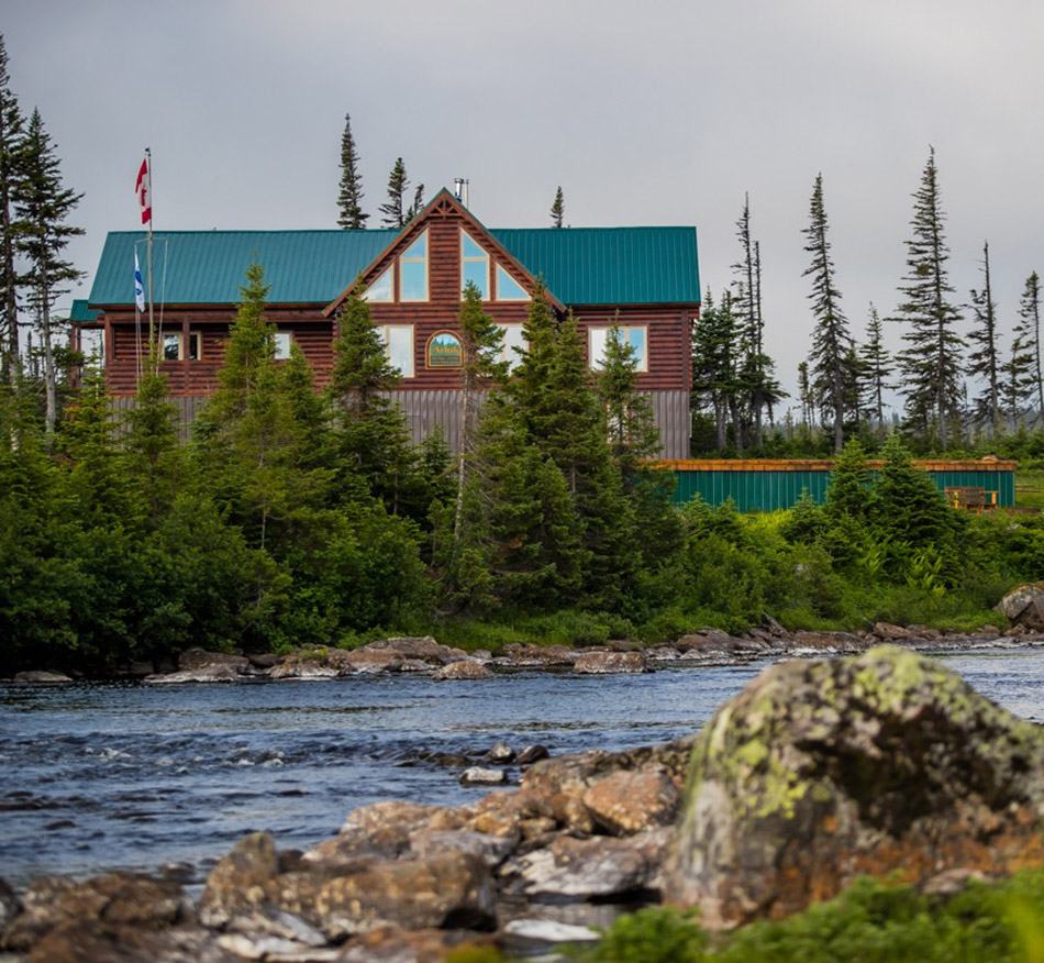 A lodge with a green roof and a flag stands behind spruce above a rocky river.
