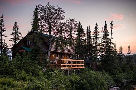A log lodge with a wraparound deck stands among tall spruce at sunset, its windows glowing.