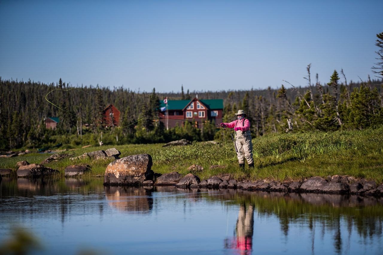 Red lodge buildings with green roofs stand beside a calm river at dusk, with an angler casting from the bank.