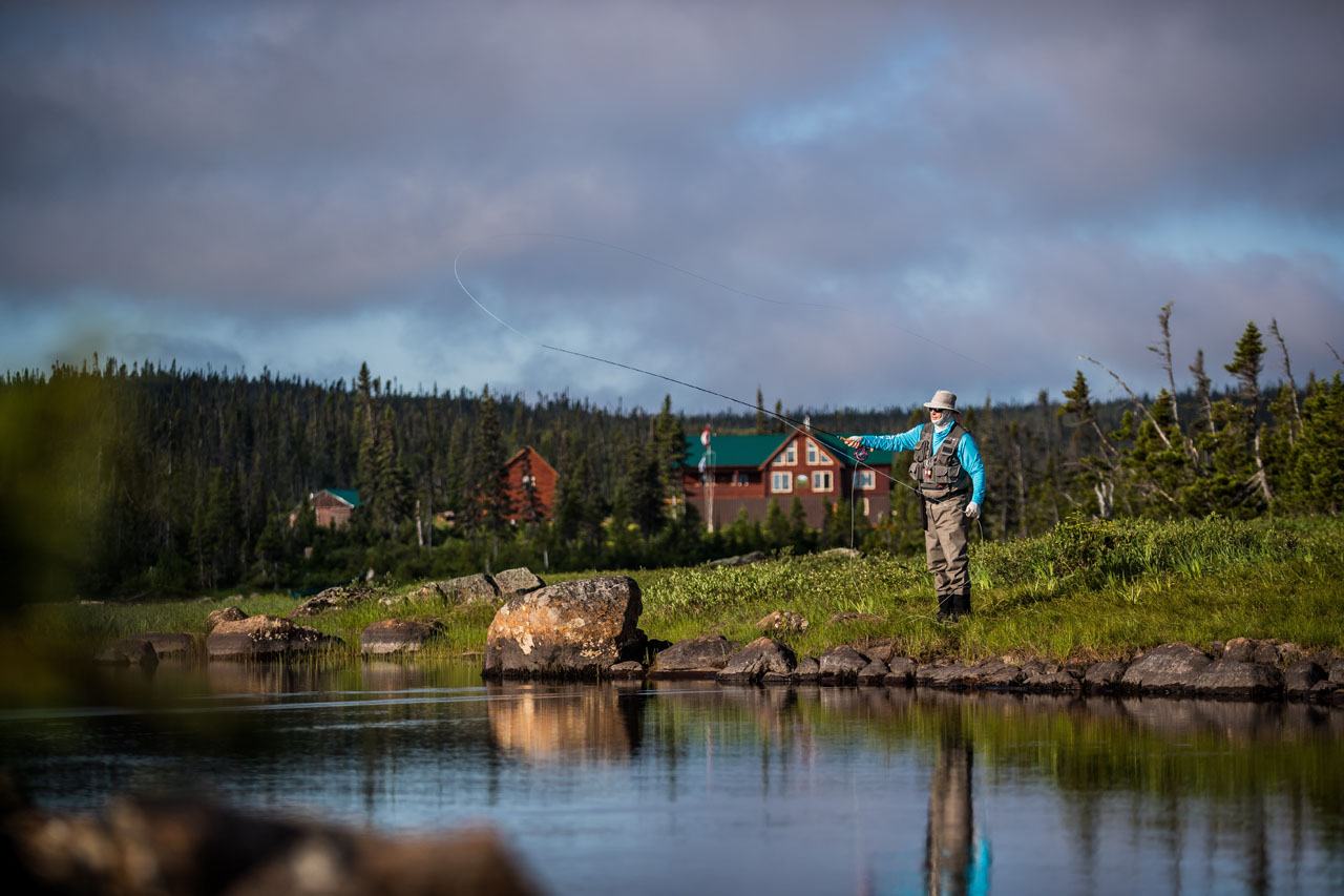 A lodge with green roofs sits on a green bank across a calm river as an angler casts in the evening light.