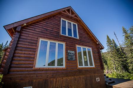 A log lodge with a steep roof and large windows stands among spruce on a bright summer day.