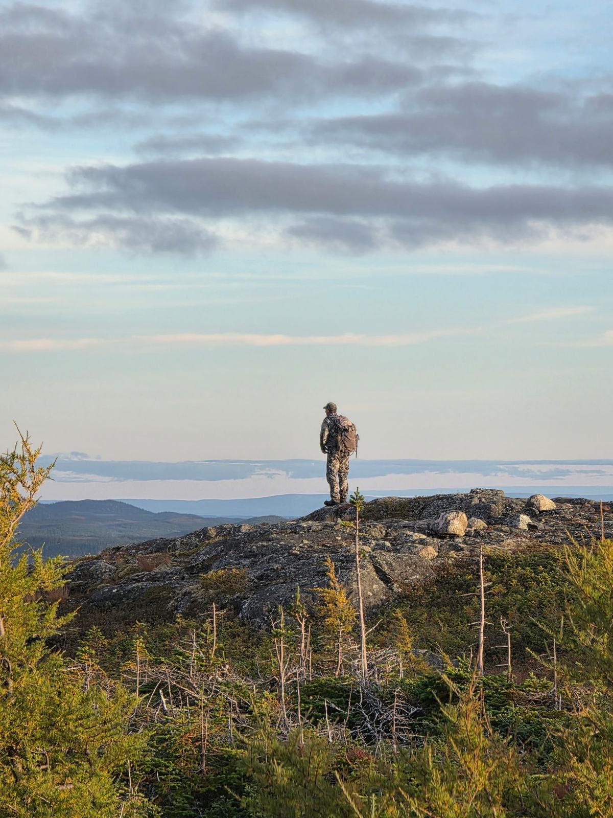 A hunter stands on a rocky ridge in evening light, with mist filling the valley below.